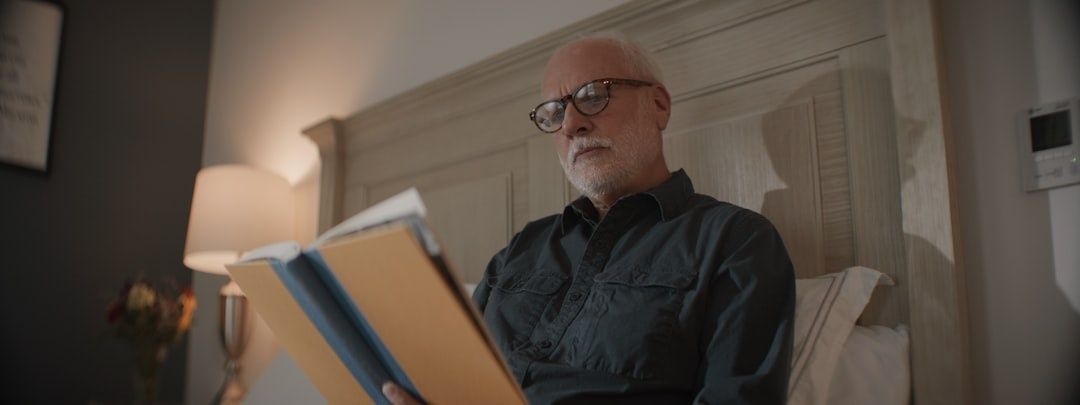 Äldre man med glasögon läser en bok i ett varmt belyst sovrum